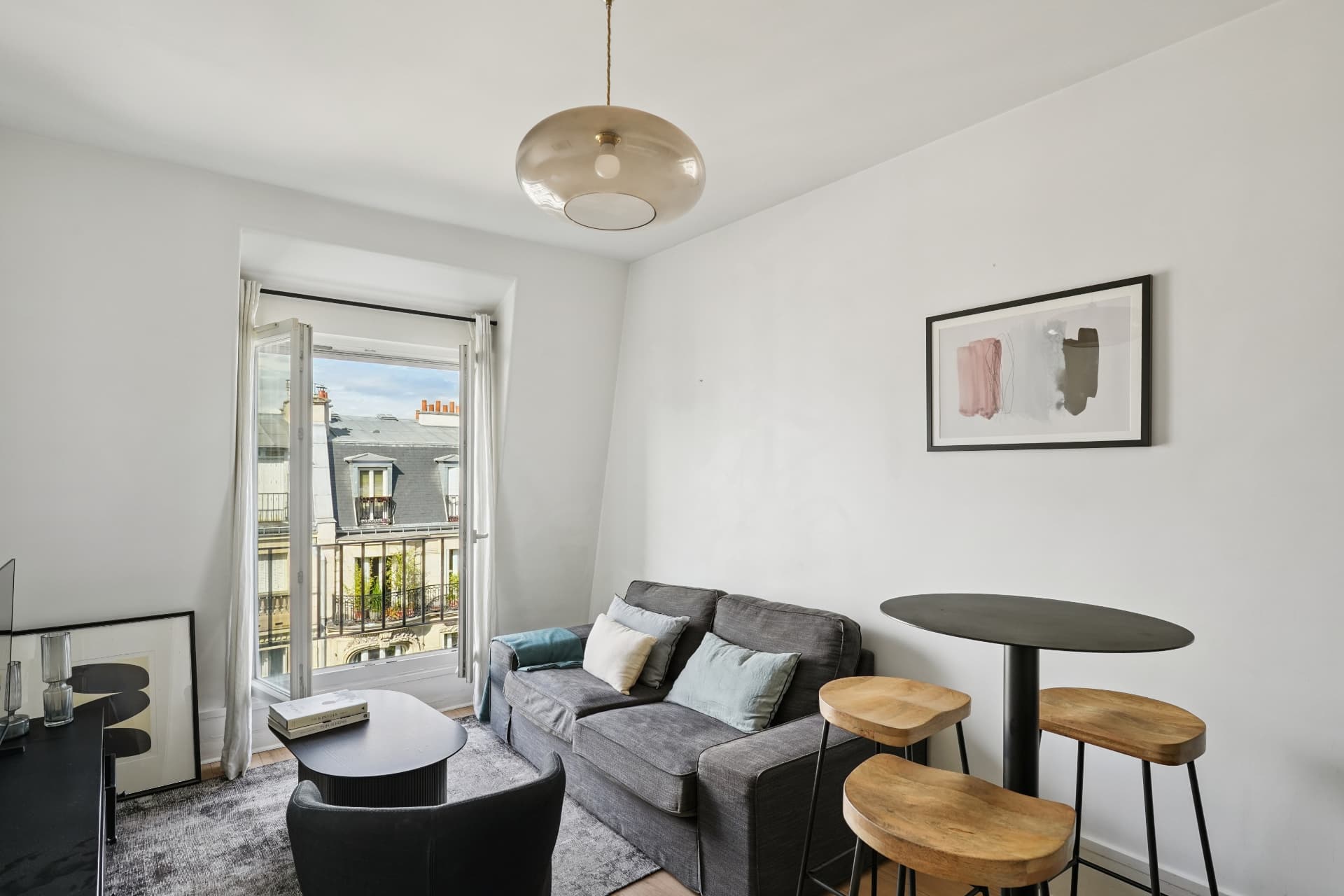 Séjour du lot 19 avec canapé gris, table haute en bois et vue sur les toits depuis le balcon