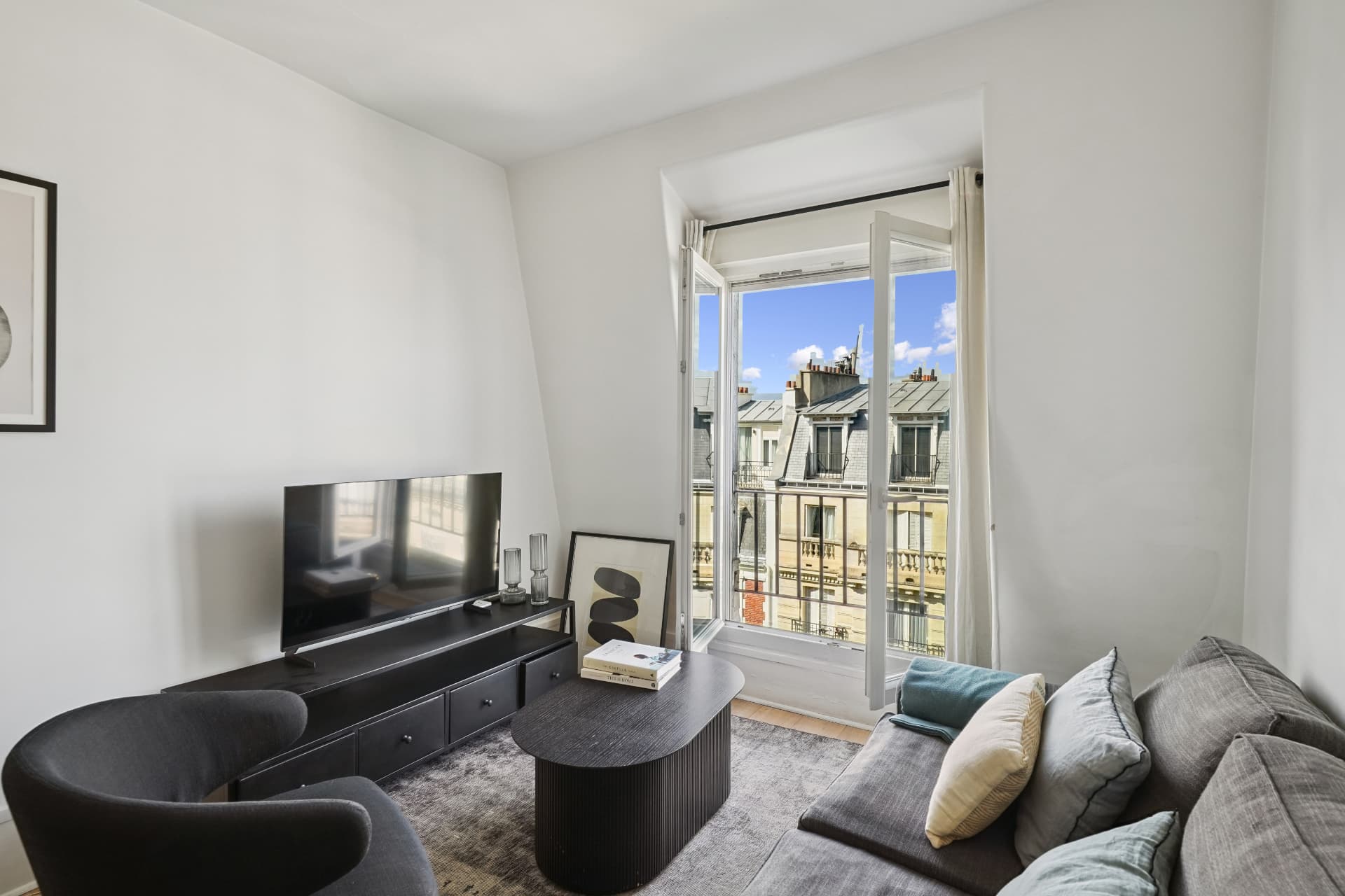 Séjour du lot 19 avec télévision, fauteuil gris et porte-fenêtre ouverte sur les toits