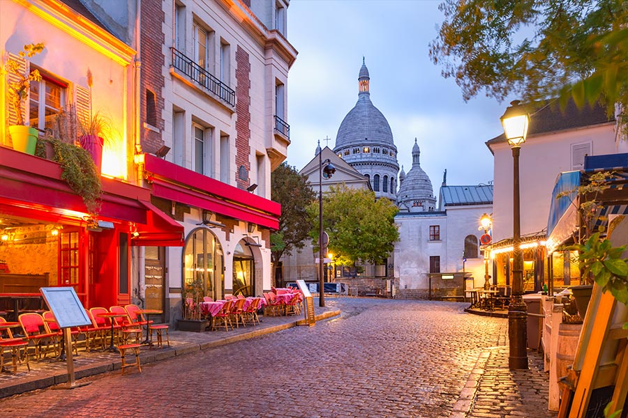 Rue pavée de Montmartre avec terrasses de café et dôme du Sacré-Cœur au loin